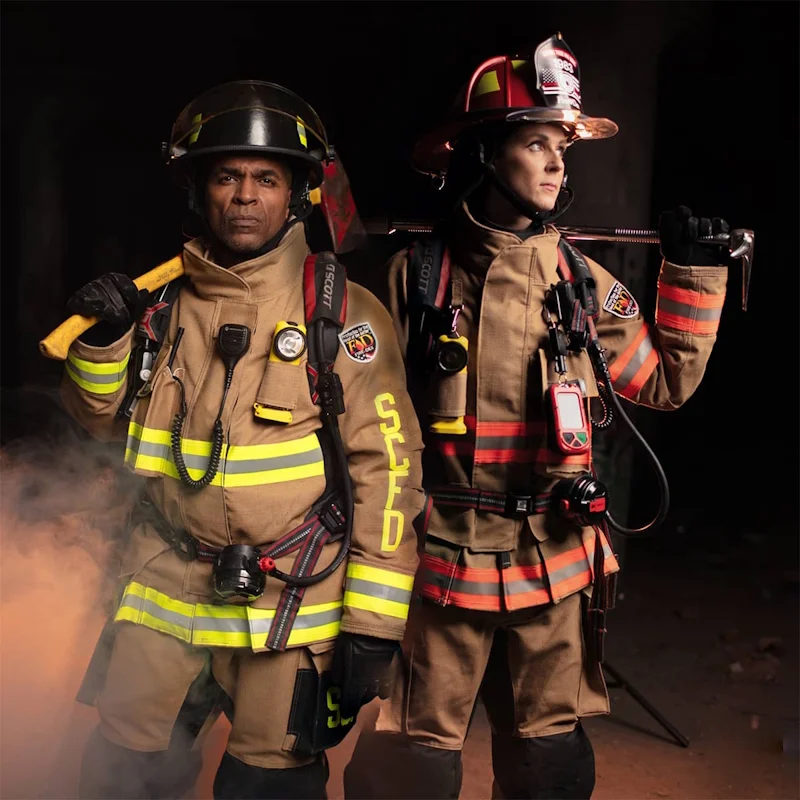 Two firefighters wearing black Fire-Dex AeroFlex turnout gear fully equipped with helmets, gloves, and tools standing ready for emergency response.