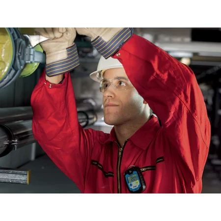 Worker in a red coverall, hard hat, and safety glasses wearing a Dräger Pac® 6000 Single-Sensor Gas Detector on the chest while working on industrial equipment.