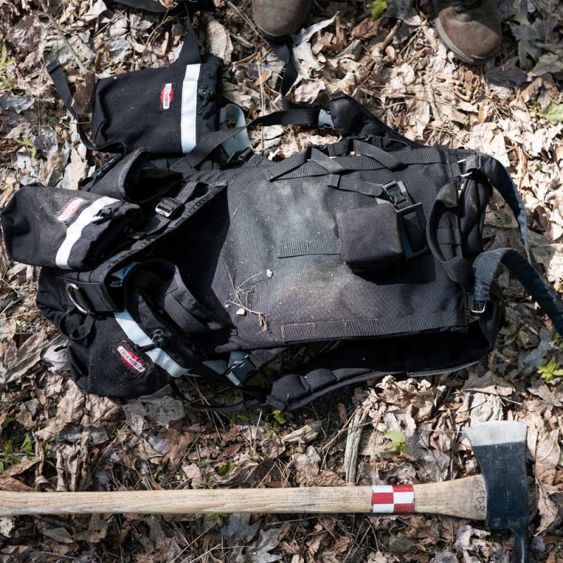 True North Gear Chainsaw Pack laying on the ground beside an axe, showcasing durable construction and ready-for-use firefighting tools.