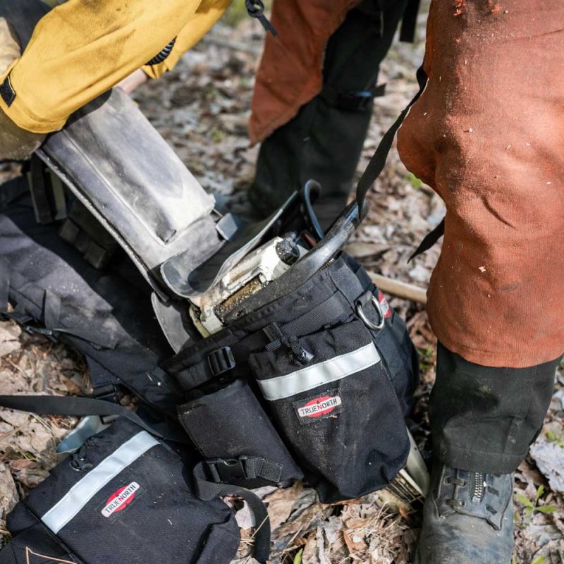 Close-up of True North Gear Chainsaw Pack placed on the ground with open compartments and chainsaw partially removed, ready for use in forestry or firefighting.
