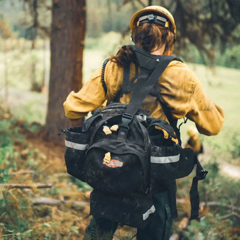 Black True North Gear Fireball™ Pack worn by a firefighter navigating through wooded terrain, highlighting its rugged durability and functional design for efficient firefighting and rescue in remote outdoor environments.