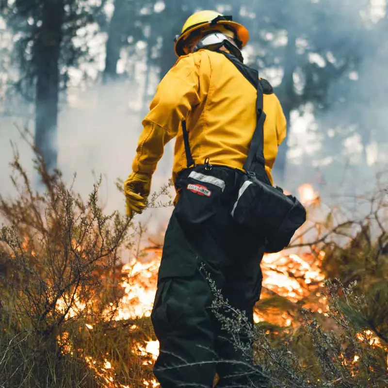 Firefighter wearing the True North Gear Spyder™ Gear Pack while navigating through a forest fire, showcasing the pack’s rugged durability and ergonomic design for carrying essential gear in challenging wildfire conditions.