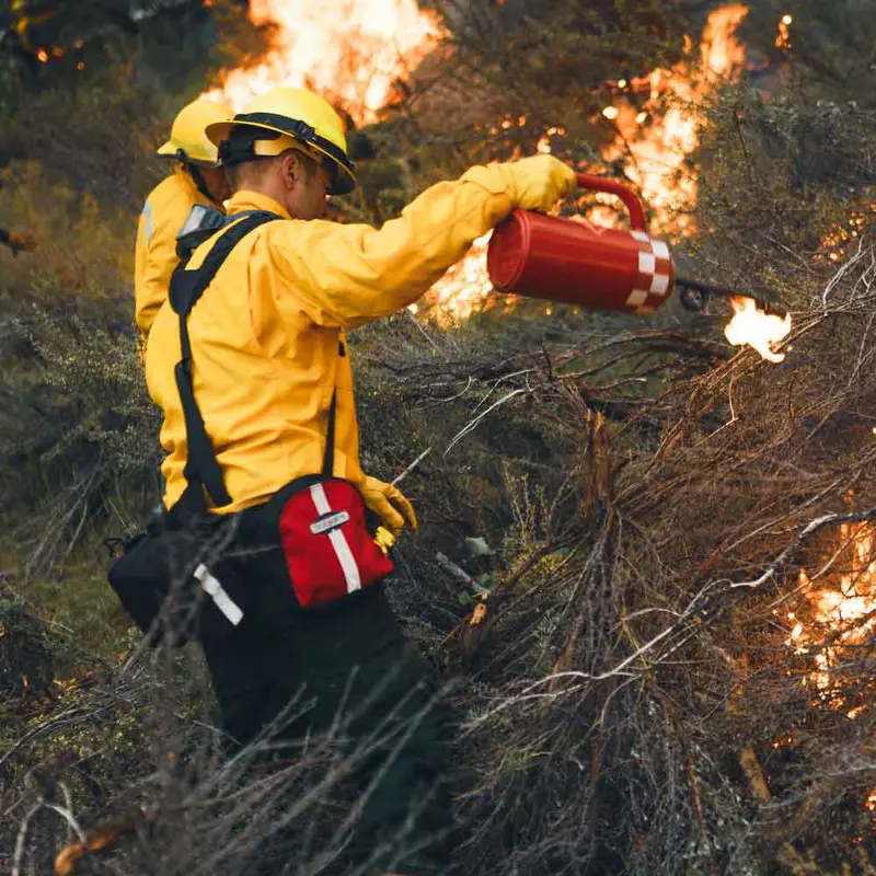 Firefighter wearing the True North Gear Spyder™ Gear Pack, cautiously navigating through active flames and smoke, highlighting the pack’s durable build and ergonomic fit for essential gear transport in hazardous wildfire conditions.