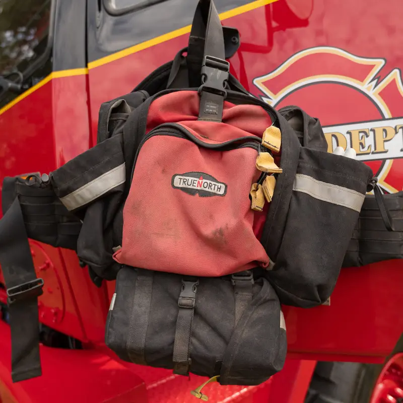 Red True North Gear Fireball™ Pack resting on a fire truck door, highlighting its rugged construction and easy access design tailored for firefighting and rescue operations.