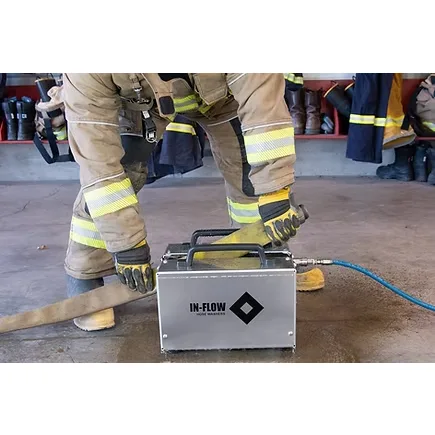 Firefighter in turnout gear feeding a fire hose through an OTEX In-Flow Hose Washer connected to a blue water supply hose.