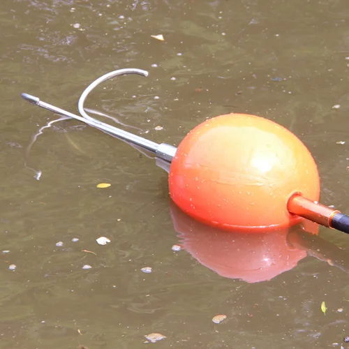 Northern Diver Inflatable Buoy Reach Pole Attachment fitted with grapple hook on a reach pole, floating on the surface of murky water for water rescue use.