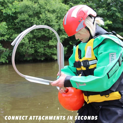 Rescuer in green drysuit and red helmet attaching a Northern Diver water rescue jaws attachment to a reach pole with buoy near a river.