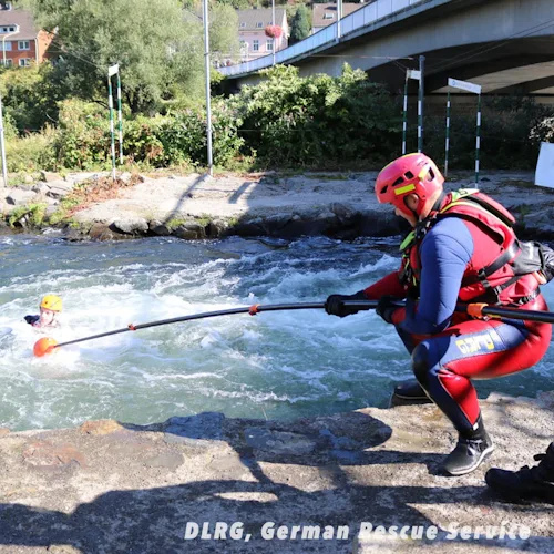DLRG German Rescue Service diver extending a Northern Diver reach pole to a swimmer in fast-moving whitewater near a bridge.