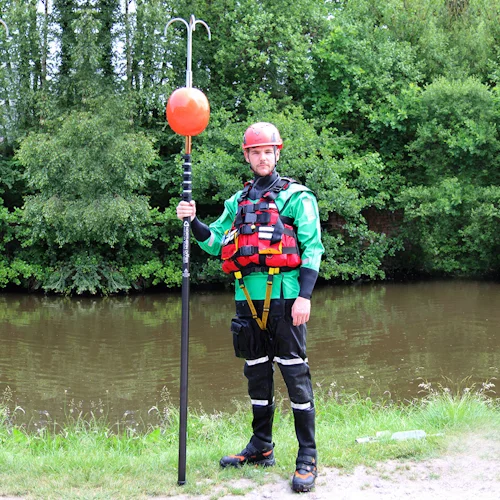 Rescuer in helmet and drysuit standing by water holding a Northern Diver reach pole fitted with a grapple hook and buoy attachment.