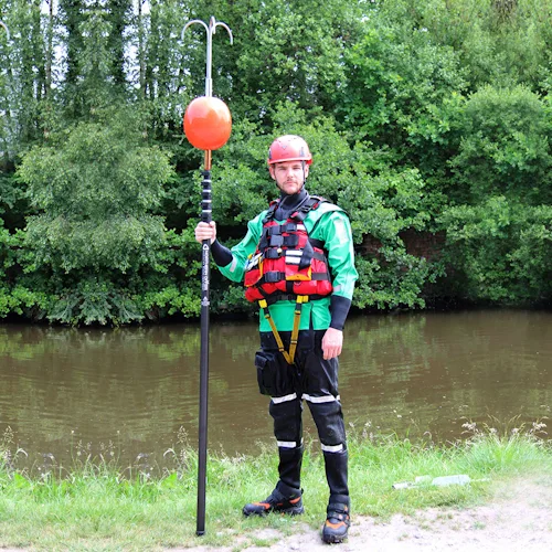 Rescuer in helmet and drysuit standing by water holding a Northern Diver reach pole fitted with a grapple hook and buoy attachment.